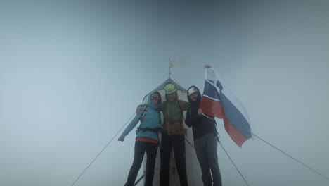 hikers on the top of a mountain triglav next to the aljaž tower with a slovenian flag happy looking at the camera