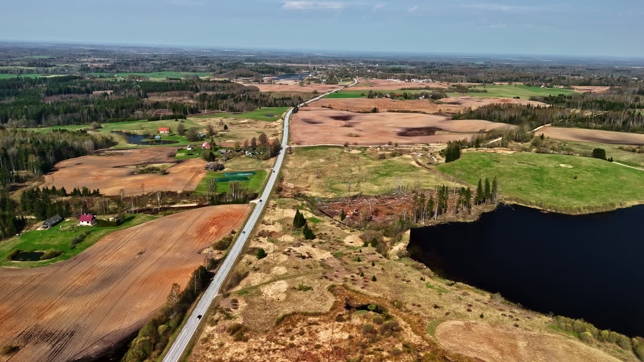 Dynamic aerial hyperlapse flying forward over a long rural road, patchwork fields, forests, and lakes in the Latvian countryside