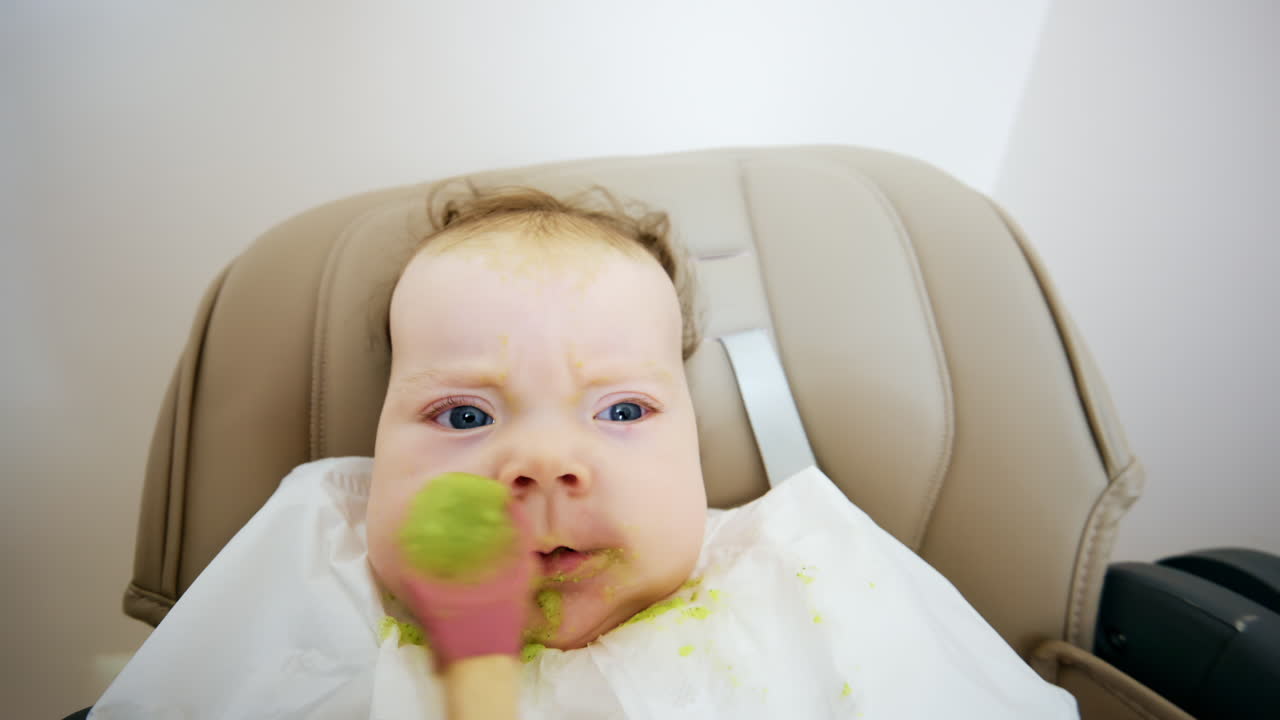 Close up of a baby eating green vegetable puree from a spoon while sitting in a high chair