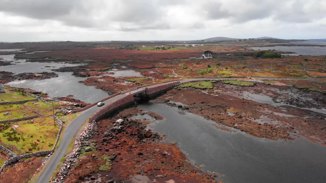 Un'auto argentata attraversa un vecchio ponte di pietra sopra un'insenatura tidale nel paesaggio aspro e paludoso del Connemara, Contea di Galway, Irlanda.