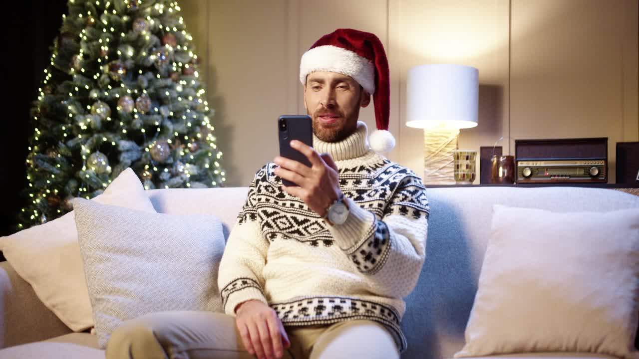 hombre guapo alegre con sombrero de santa sentado en una habitación acogedora con árbol de navidad y chateando por video en un teléfono inteligente felicitando con las vacaciones
