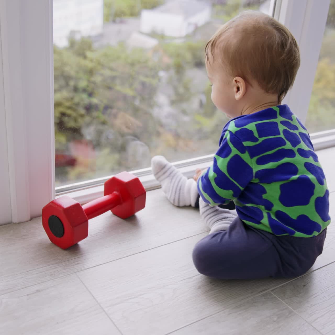 Little adorable baby boy playing with a red dumb-bell on the floor. Lovely toddler crawling by the floor at big panoramic window. High angle view