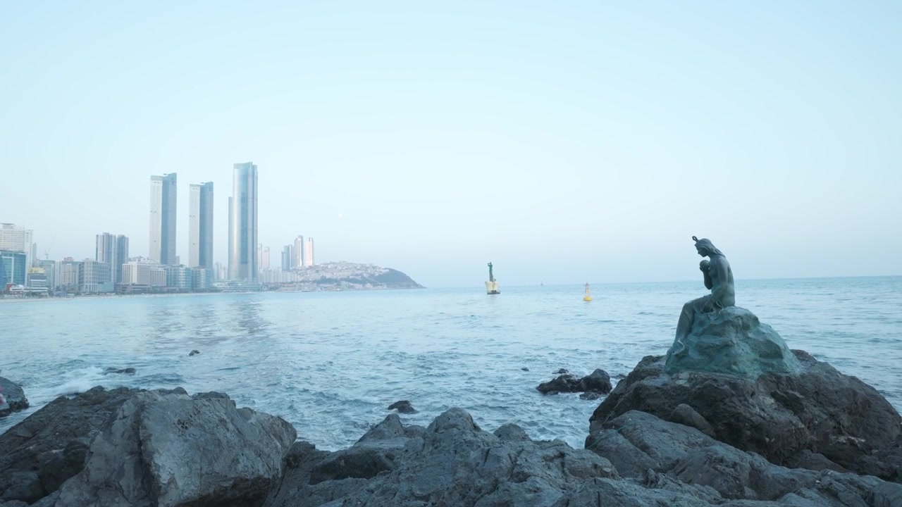 La statua della sirena Principessa Hwangok si trova sulle rocce costiere che si affacciano sullo skyline della città vicino alla spiaggia di Haeundae a Busan, Corea del Sud. Campo lungo
