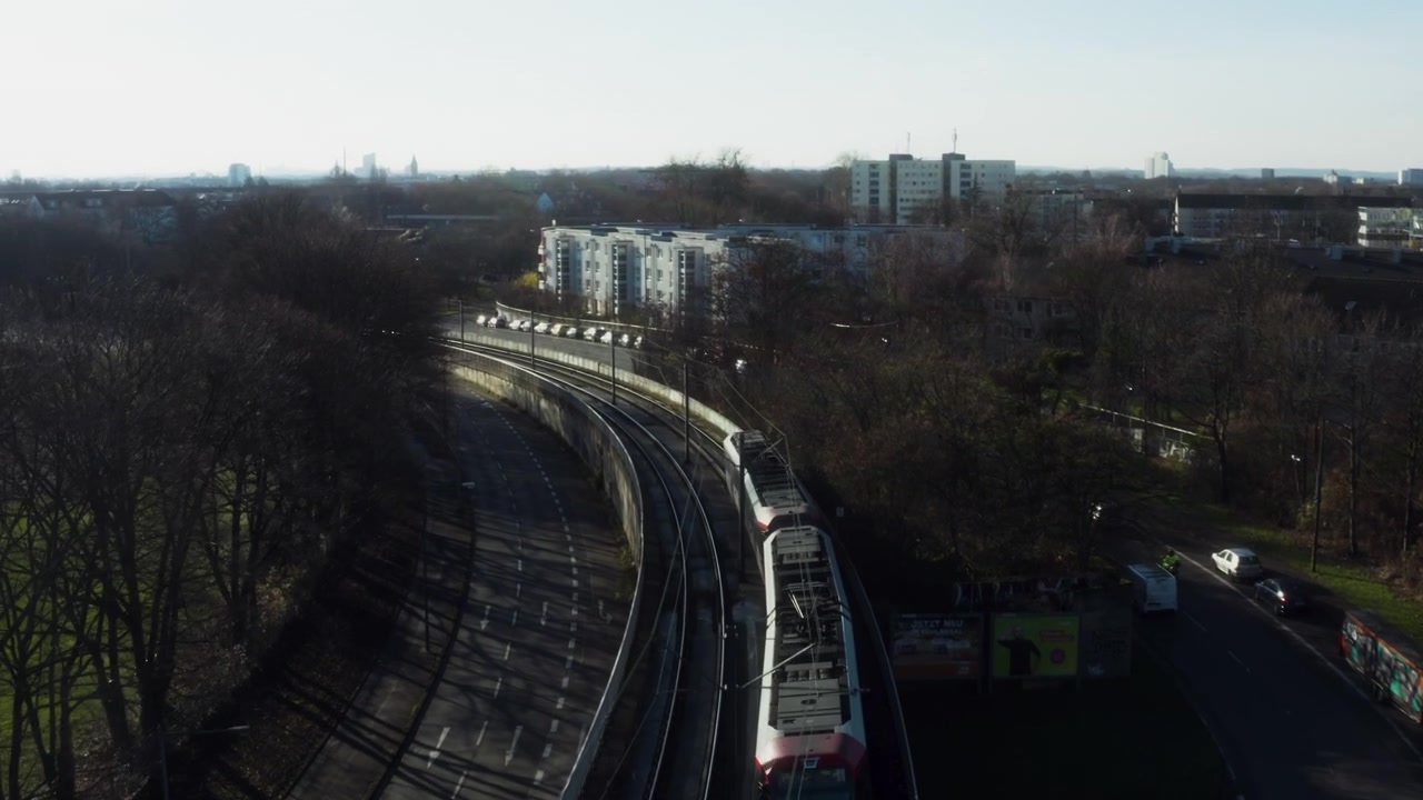 Drone ascending above a metro line in Cologne, Germany, as a commuter train moves away through the urban landscape and rail infrastructure.