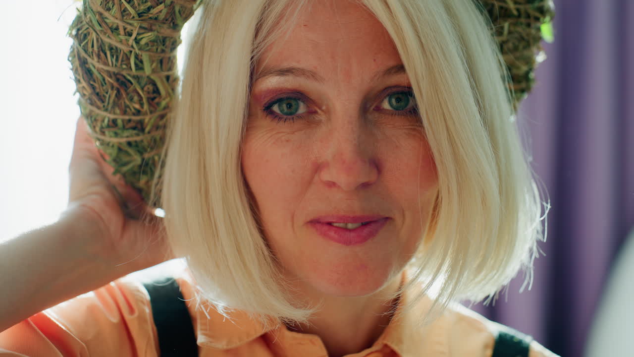 Blonde woman adjusting rustic floral wreath hat made of hay and flowers while standing in bright sunlight, soft lens flare effect highlighting natural and artistic atmosphere