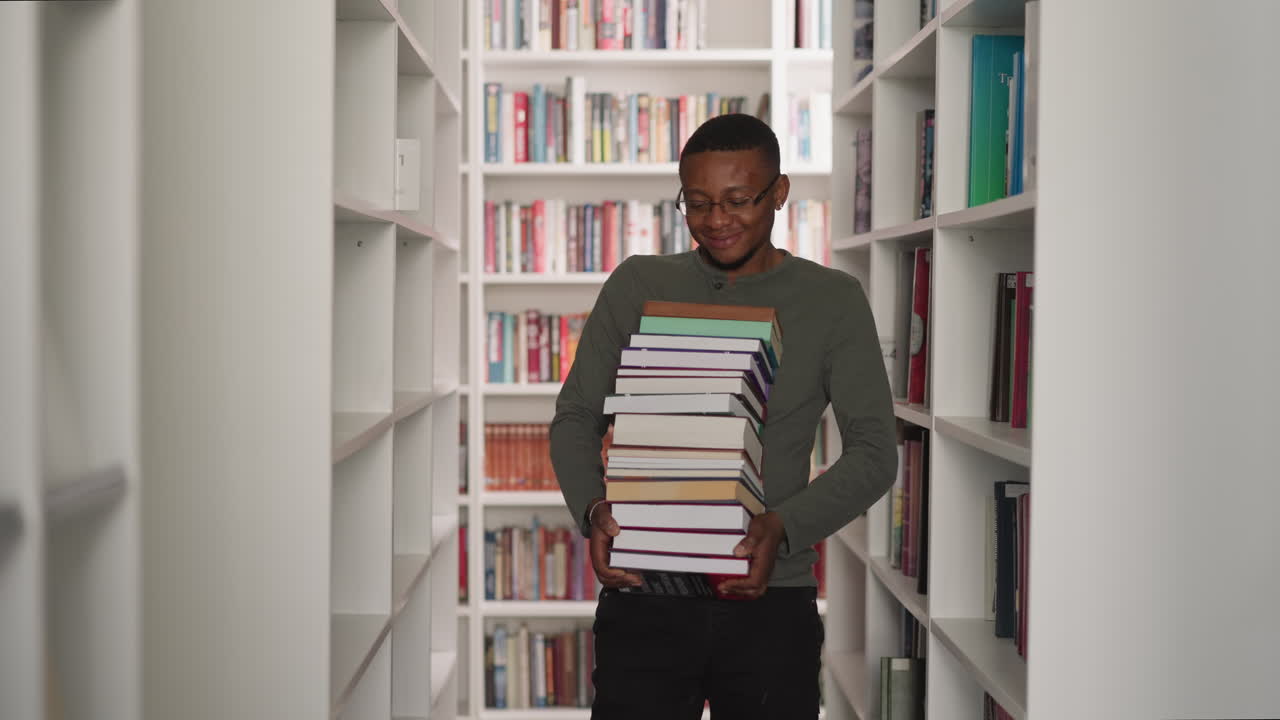 Emotional man carries book stack in library. African American librarian holds large literature pile walking between bookcases. Student in archive