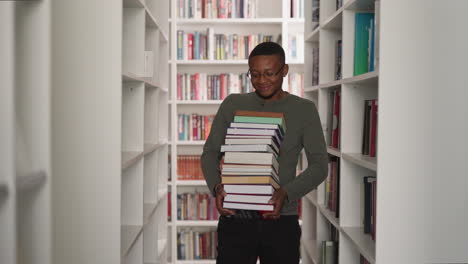 emotional man carries book stack in library. african american librarian holds large literature pile walking between bookcases. student in archive