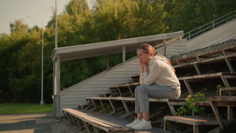 thoughtful young woman in casual attire sitting on rustic stadium bleachers with hands resting on chin, surrounded by greenery in peaceful outdoor setting, she appears pensive and reflective