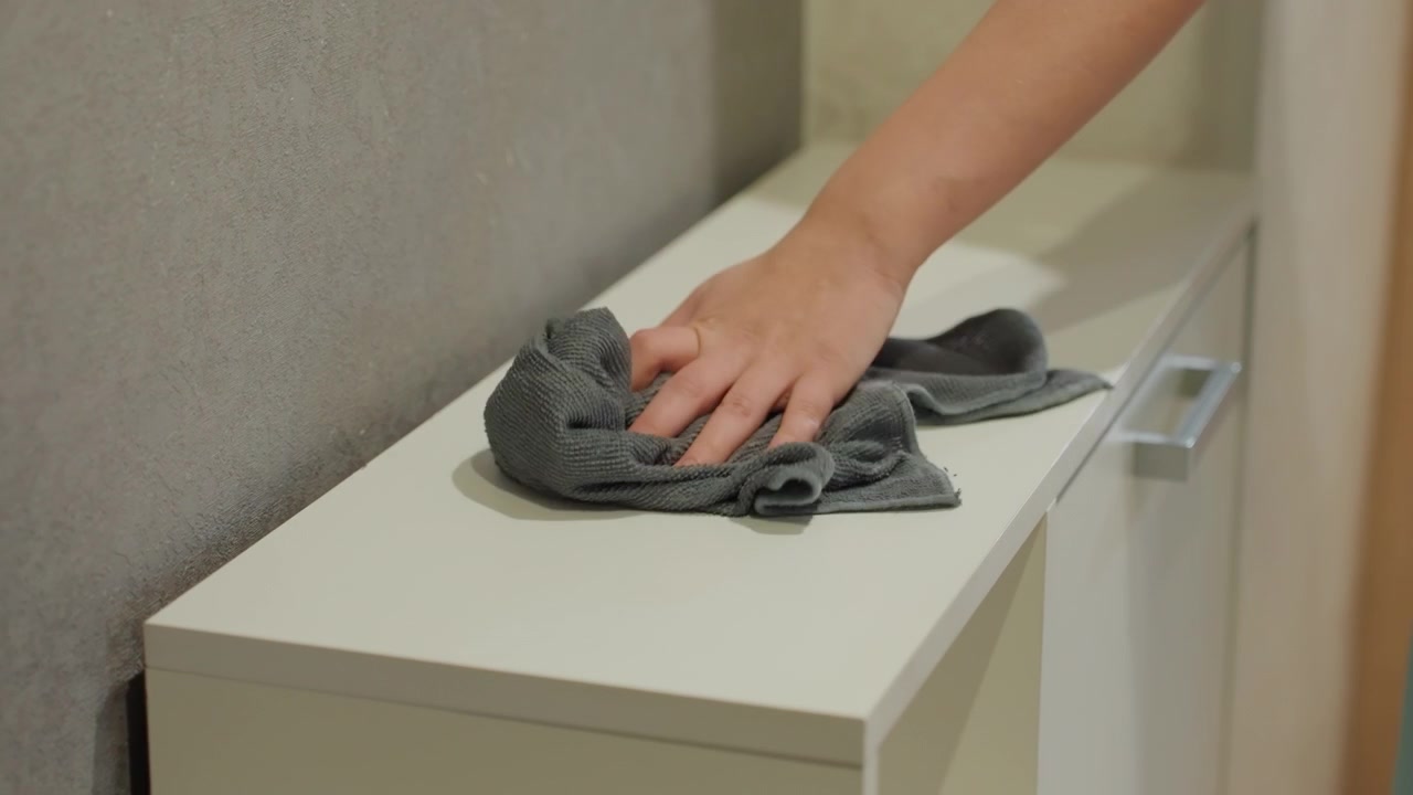Detail rukou utírajících horní část skříňky utěrkou z mikrovlákna, soustředěný pohyb, texturovaná stěna, bílý lakovaný povrch, jemný tlak, pečlivé čištění, záměrná detailní práce a uspokojivý výsledek.