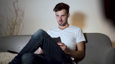 a young man with a stubble and a modern hairstyle, wearing a white t-shirt and jeans, sitting bored at home on a sofa, legs crossed, smiling while browsing his tablet, static 4k