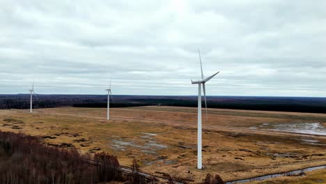 Gruppe-Von-Hohen-Weißen-Windrädern-Mit-Drei-Langen-Rotorblättern,-Die-Auf-Einem-Riesigen-Grünen-Feld-Unter-Einem-Bewölkten-Himmel-Stehen,-Zeitlupe,-Luftaufnahme