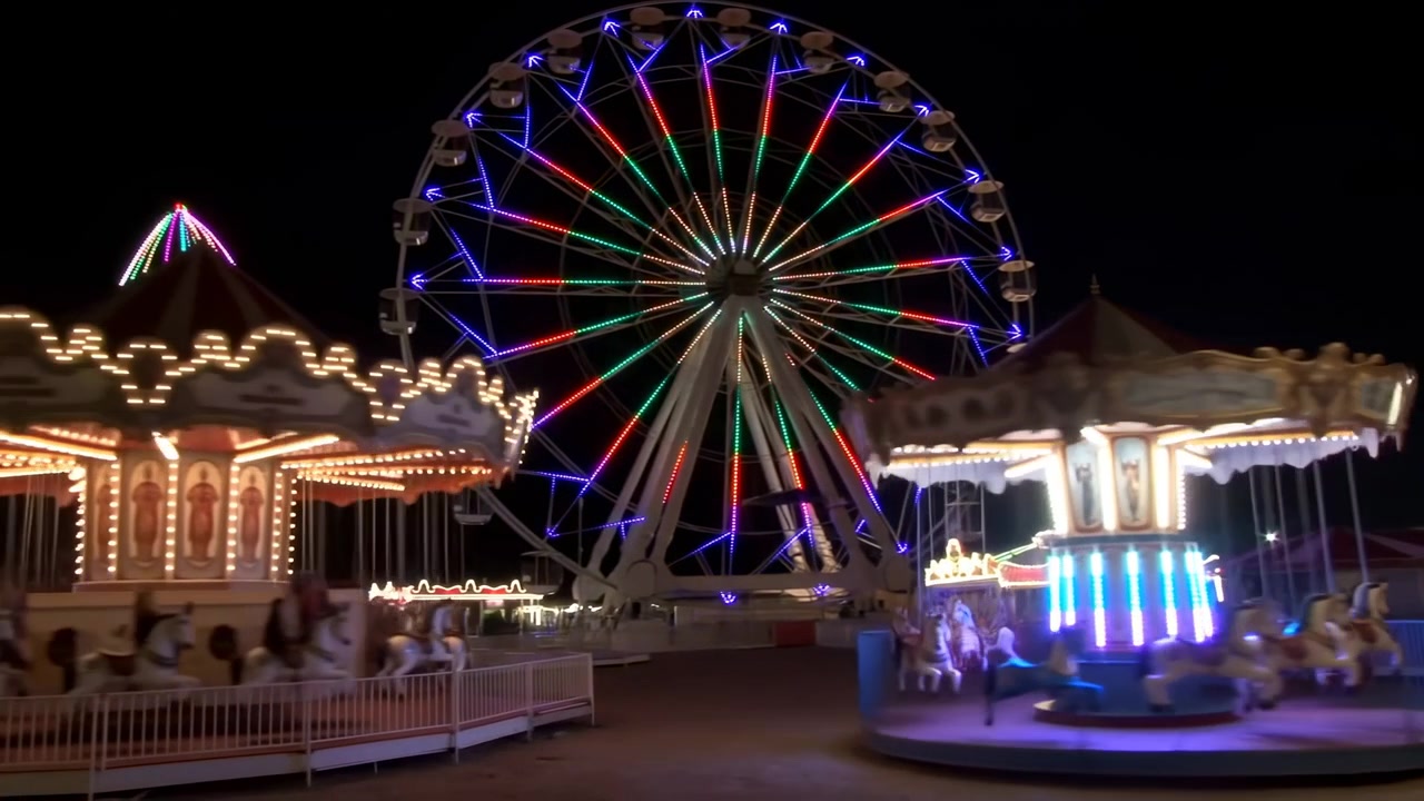Rotating carousel with blurred horses and vertical light columns spins in foreground while illuminated ferriswheel with multicolored spokes and rim lights glows behind against dark sky