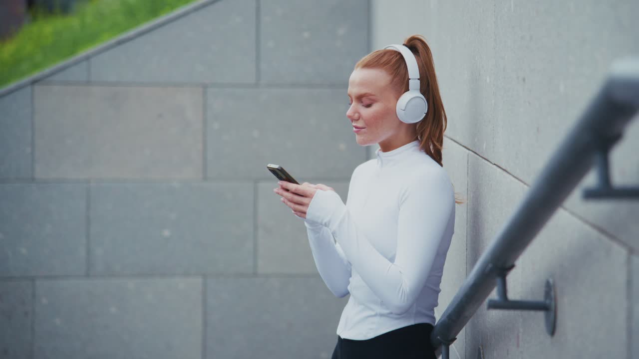 Redhead woman with headphones using phone by urban wall