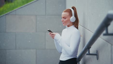 redhead woman with headphones using phone by urban wall