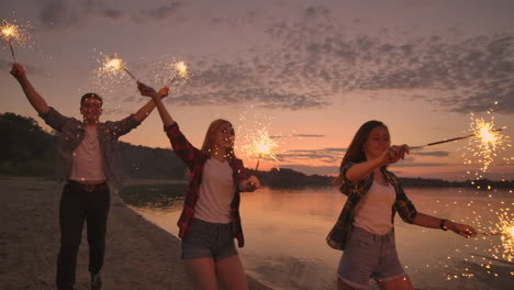 alegres amigos masculinos y femeninos están corriendo a lo largo de la playa al atardecer sosteniendo fuegos artificiales brillantes y luces fugitivas en cámara lenta. baile y fiesta de atardecer en la playa
