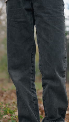 vista de ángulo bajo de una persona con pantalones de jean de pie en el bosque mientras vierte hojas secas de una bolsa en el suelo, con follaje de otoño esparcido en el fondo durante la temporada de otoño