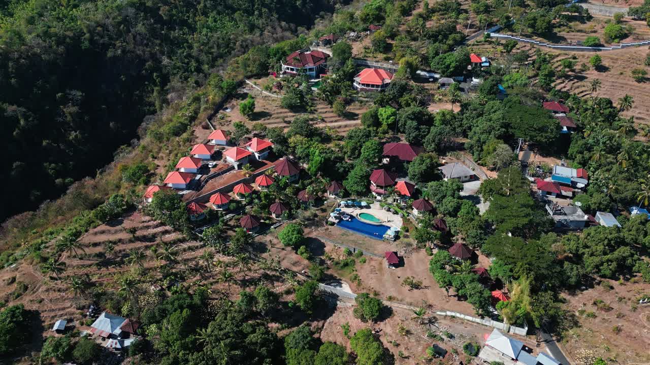 Imagem aérea de drone de uma vila balinesa, mostrando casas tradicionais cercadas por árvores verdes densas, capturando uma atmosfera tropical quente e vibrante.