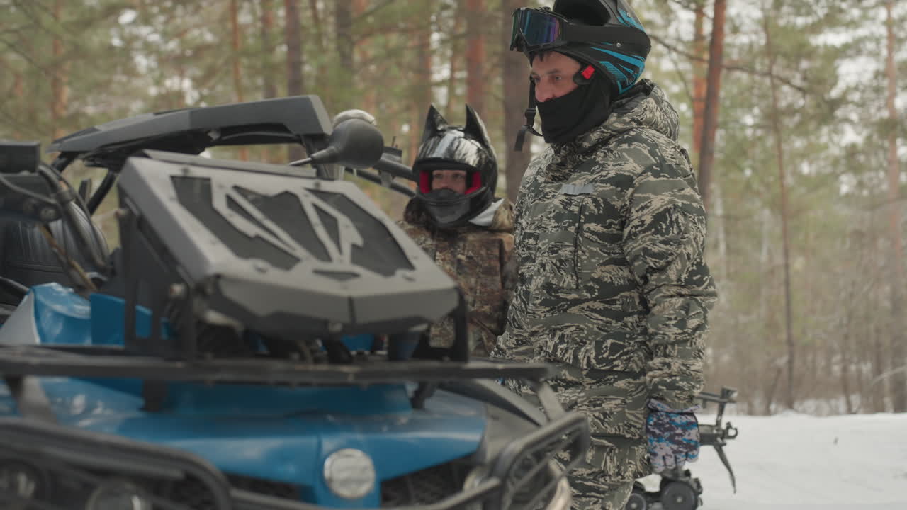 Un pilote de quad en forêt enneigée inspecte son quad, veste de camouflage et casque avec GoPro, quad bleu garé sur la neige parmi les pins, respiration visible dans l'air froid, gants mis, coéquipier en arrière-plan préparant son équipement