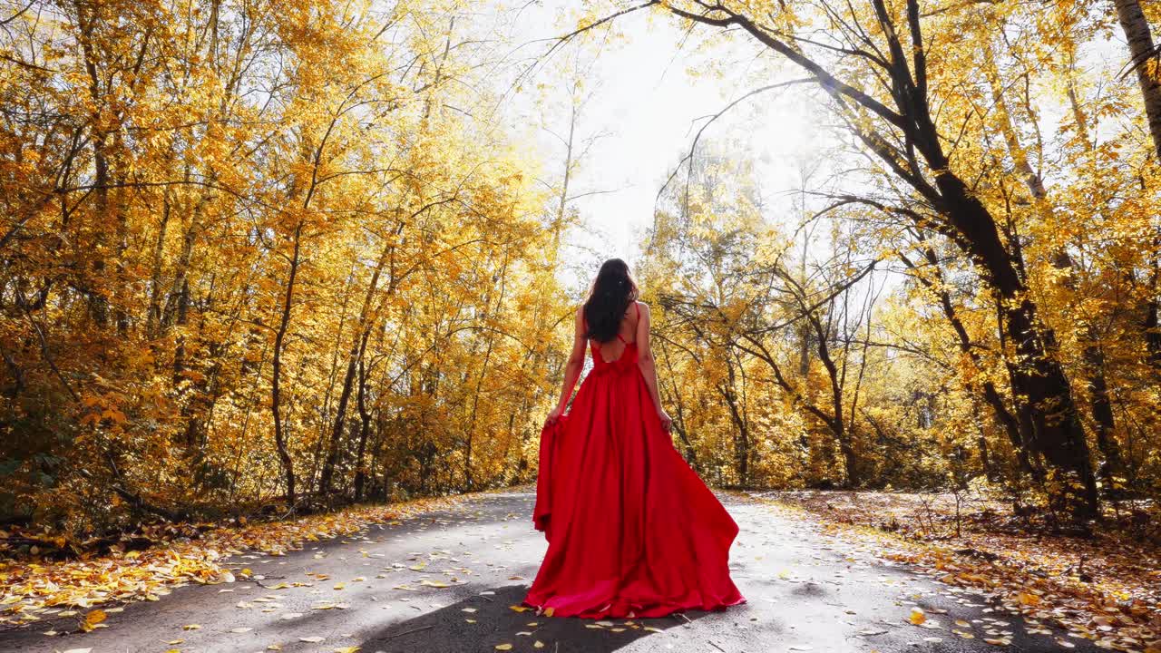 A Stunning Portrait of Elegance Amidst Autumn's Splendor: A Woman in a Flowing Red Dress Stands on a Leaf-Covered Path Surrounded by Golden Trees