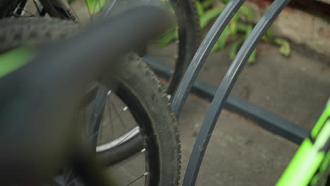 close-up view of bicycle wheel parked in bike rack alongside other bicycles in background, focus on wheel, spokes, and metal stand with blurred background elements