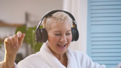 cheerful senior woman listening to music in headphones and dancing near window
