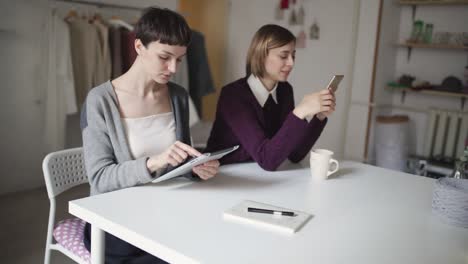 Two-woman-students-using-tablet-and-mobile-phone-sitting-at-table