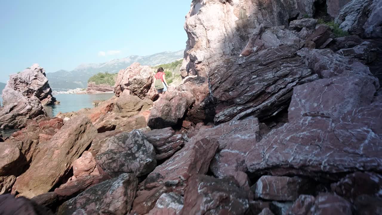 Lady walking over the rocky coastline with a really calm waters on the adriatic coastline in Montenegro. Wearing a red top and denim shorts whilst the camera follows her
