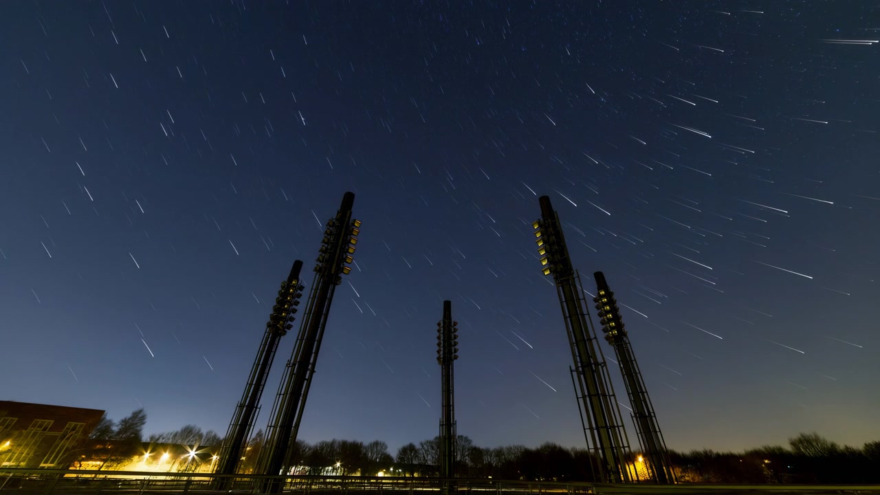 Low-angle panoramic night scene showing several stadium floodlight towers in dark silhouette while bright star trails arc across indigo sky, creating dramatic contrast, open space, and calm urban mood