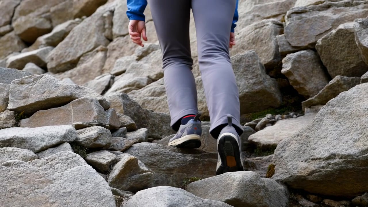 Low rear three quarter view shows lower body ascending rough stone terrain, with fitted gray pants, athletic hiking shoes, bent knee, lifted heel, and weight shifting forward during climb