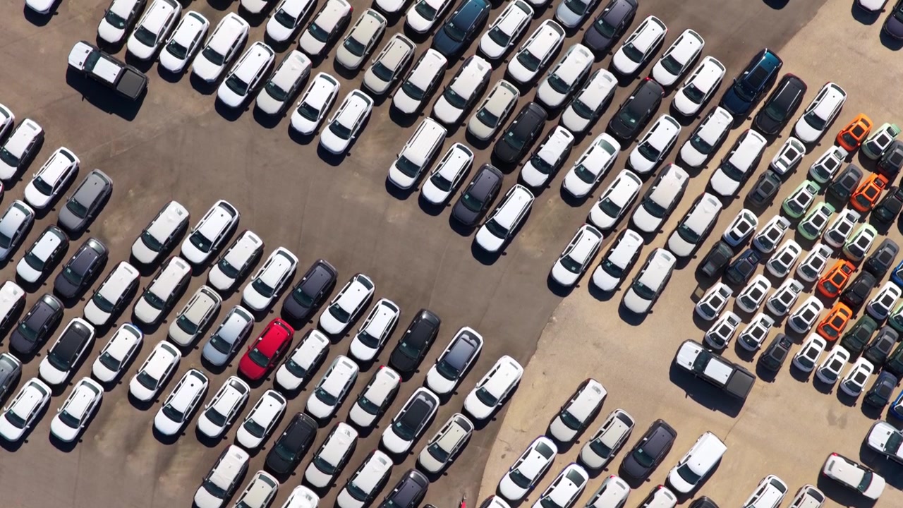 Luftfoto ovenfra viser rækker af nye biler parkeret på en stor parkeringsplads ved en fabrik eller havn, afventer salg, import eller eksport i bilindustrien og repræsenterer global handel og forsyningskæde.
