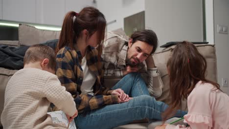 Un-Hombre-Moreno-Feliz-Con-Barba-Incipiente-Que-Viste-Una-Camisa-Beige-Se-Comunica-Con-Su-Pequeña-Hija-Y-Su-Hijo-Junto-Con-Su-Esposa-Mientras-Los-Niños-Dibujan-En-El-Piso-Cerca-Del-Sofá-En-Un-Apartamento-Moderno