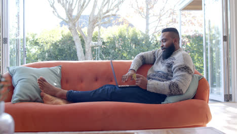 African-american-man-using-laptop-and-credit-card-in-sunny-living-room,-slow-motion