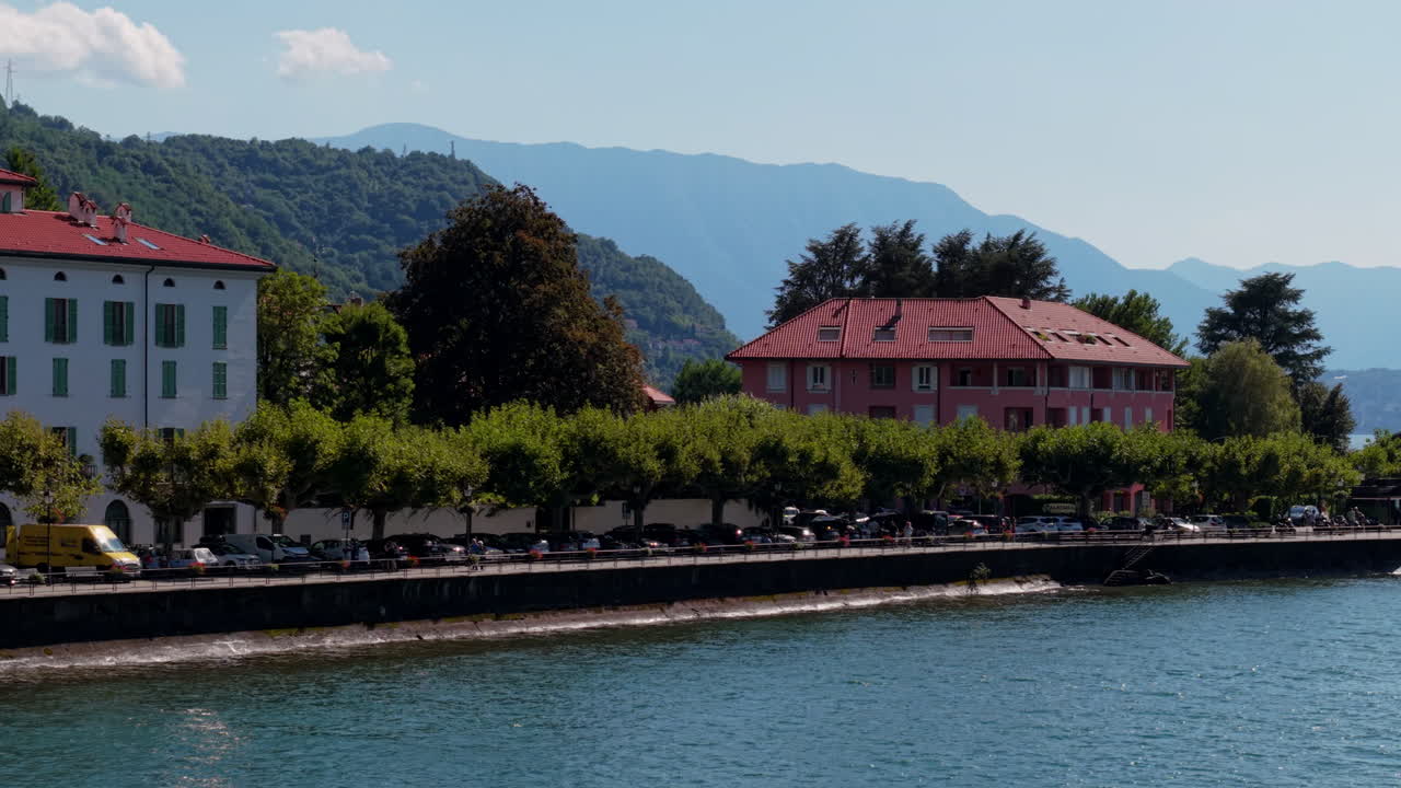 Drone rises from lakeside level to reveal rooftops, shoreline trees, and the wide expanse of Lake Como with layered mountains in bright daylight
