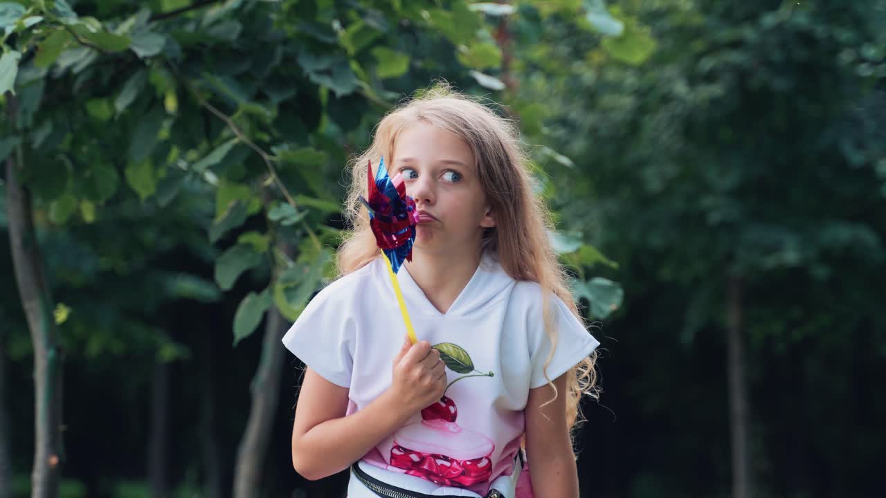 Portrait of a little mischievous girl child with a toy windmill in her hands grimaces and poses looking at the camera in a park in nature