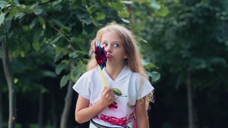 portrait of a little mischievous girl child with a toy windmill in her hands grimaces and poses looking at the camera in a park in nature