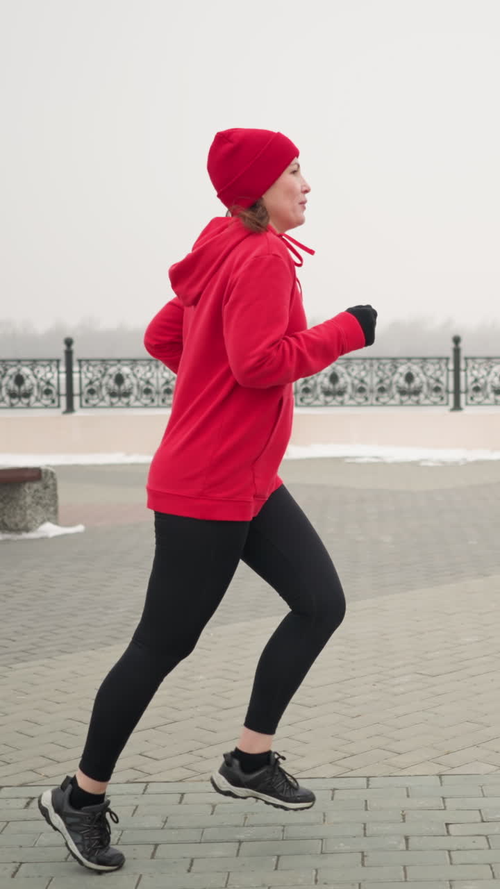 Side view of woman in red hoodie and beanie jogging outdoors during winter on interlocked pavement surrounded by serene urban elements like benches, decorative railings, and snowy surroundings