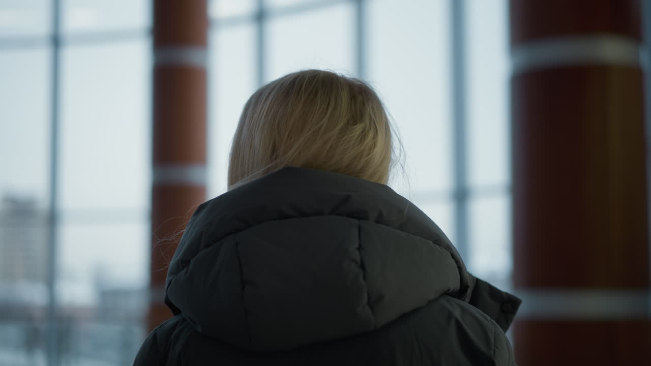 블랙 후디를 입은 여성이 현대적인 실내 공간에서 상적으로 주변을 둘러보는 클로즈업 헤드 뷰, 배경에는 도시 건물의 유리 창문이 있습니다.