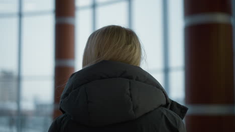 close-up head view of lady in black hoodie looking around contemplatively in modern indoor space, background features glass windows with urban buildings