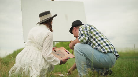 a man and woman are bent over in a grassy field, preparing for an outdoor painting session. the woman in white holds a palette, while the man in a checked shirt and hat picks up a paint tube