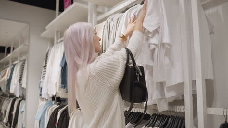 female shopper with pink hair wearing white knit turtleneck sweater browses clothing store racks, lifting polo shirt with both hands while deciding purchase during seasonal sale under bright lighting