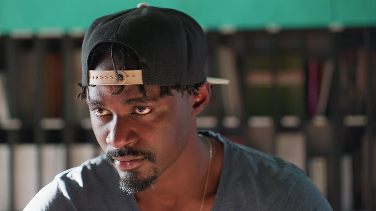 Man wearing cap sits at library table with stack of books, slowly lifting head while looking away, eyes closed, teal wall and shelves behind him bathed in soft light creating introspective mood