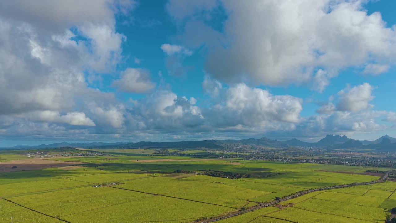 Aerial over expansive, flat agricultural land in Mauritius. The shot captures a mosaic of green and yellow crop fields leading toward a mountain backdrop under a bright, cloud-filled sky