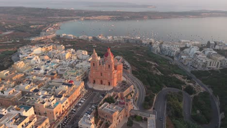An aerial view of the Mellieha Parish Church in Malta with its red dome and twin bell towers. The historic church overlooks the town of Mellieha, the scenic coastline, and the bay filled with boats