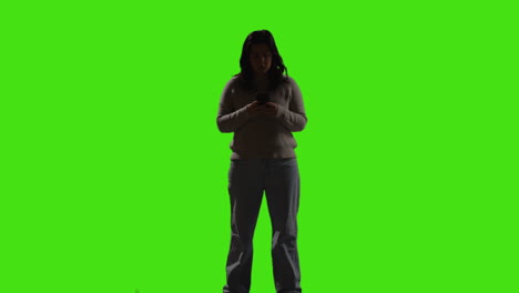 young woman sending or reading text message or scrolling online on mobile phone standing against green screen with low key lighting