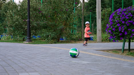 lovely baby running away from the football rolling after him. caucasian kid having fun in the park.