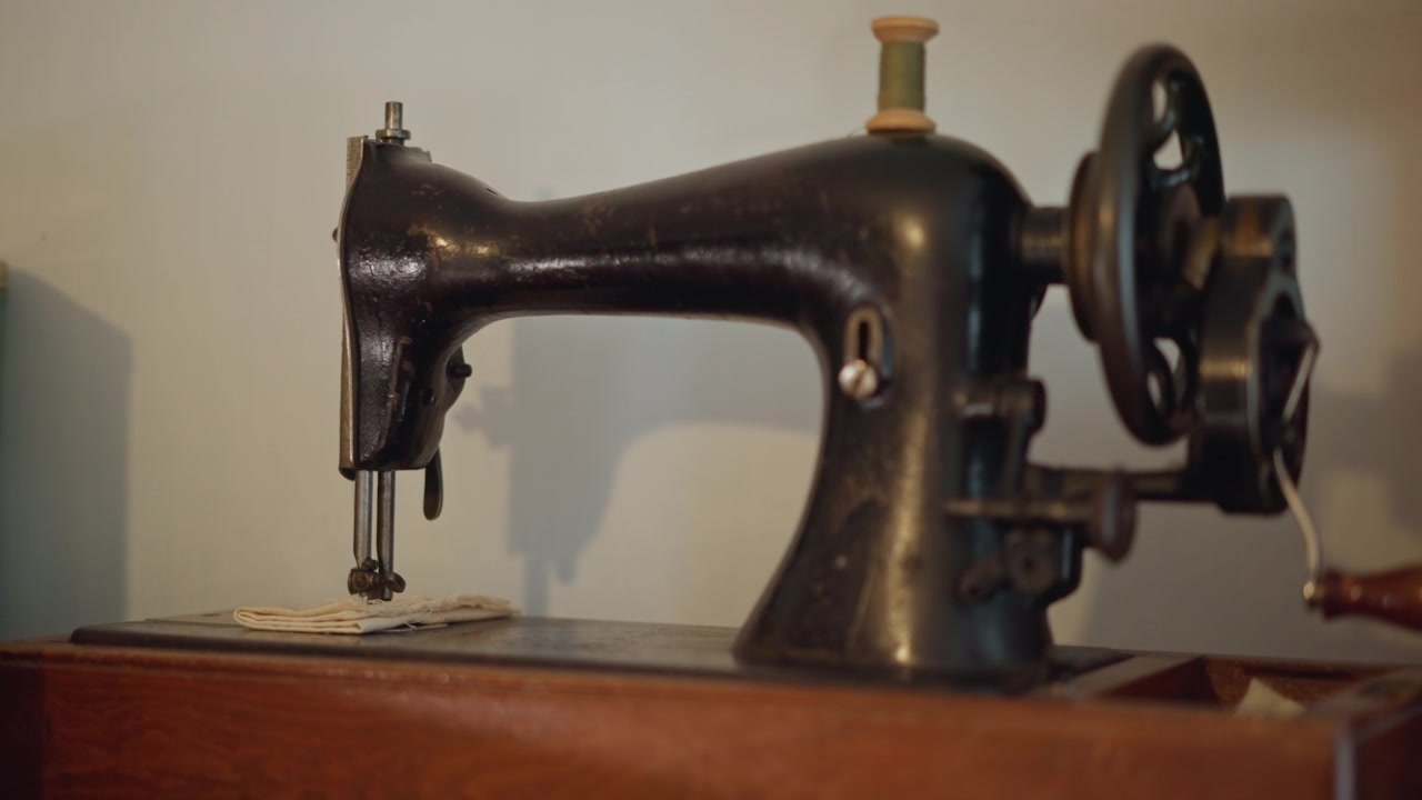 Vintage sort symaskine på bord, blødt oplyst værkstedsscene, emalje med afskallet maling, håndsving, synligt spoleområde, stofrester og metaldåse i nærheden, antydning af skrædderværktøj, varm filmkorn.