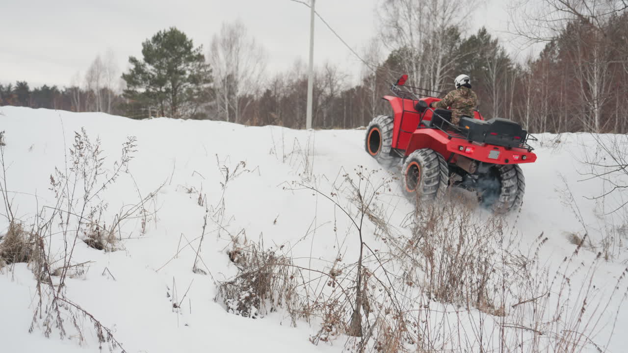 Pilota di ATV rosso in un campo innevato, con casco e giacca mimetica, guida il quad tra betulle e pini. Le gomme sollevano neve e il vapore esce dallo scarico, in un paesaggio rurale remoto di fuoristrada estremo invernale.