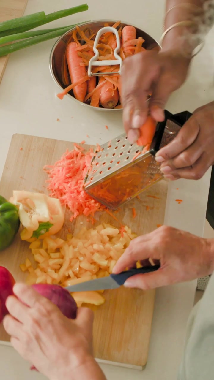 Video verticale: Una donna in grembiule color salvia grattugia una carota su una grattugia in cucina, la preparazione del pasto sta iniziando.