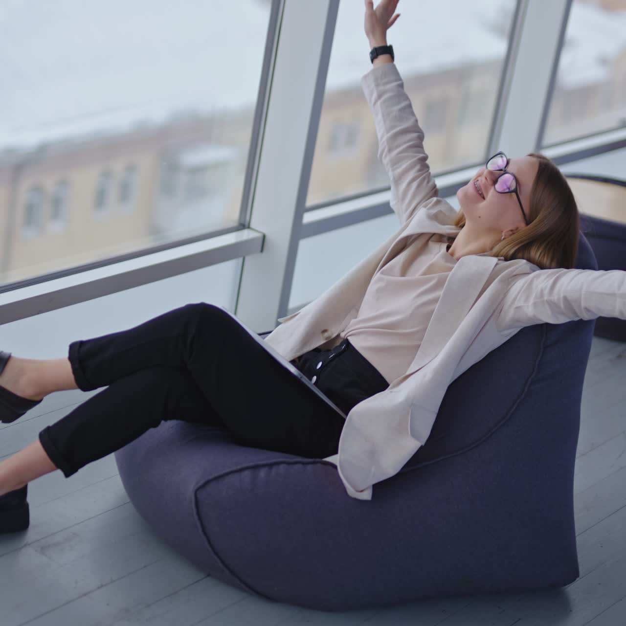 Happy relaxed woman sits in bag armchair with computer on her laps. Dreamy lady stretches hands and puts them behind her head. View from above