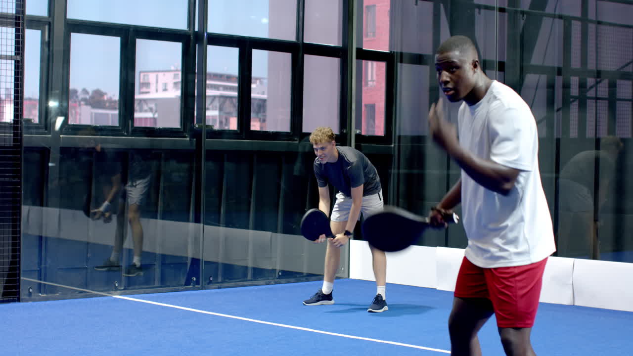 Två män med olika bakgrund spelar padeltennis på en inomhusbana och fokuserar på spelet.